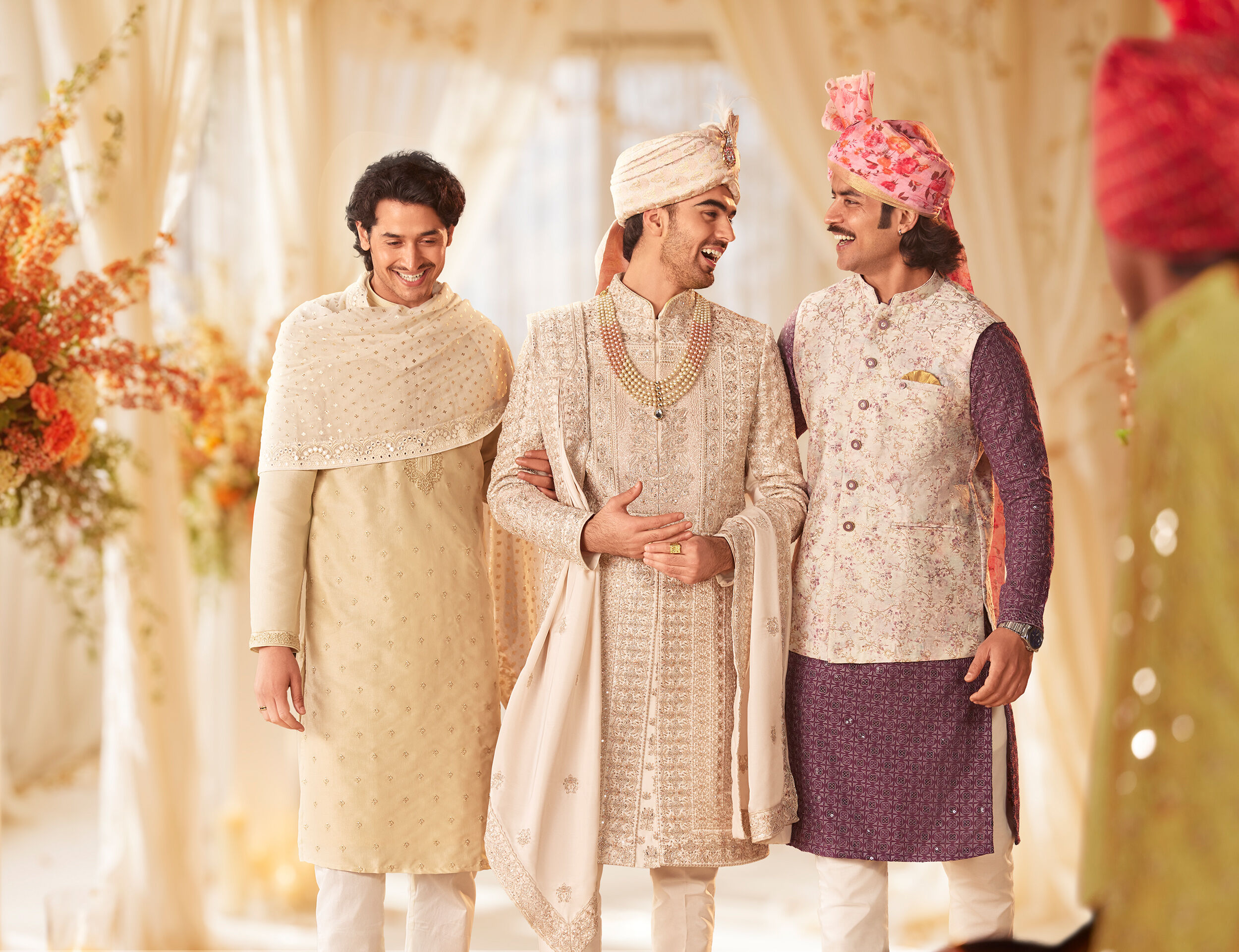 Three men in elegant Indian wedding attire, featuring a groom in a beige sherwani with turban, standing with smiling friends in traditional outfits.